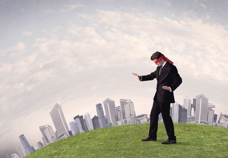 Young blindfolded businessman steps on a a patch of grass with a grey buildings in the background