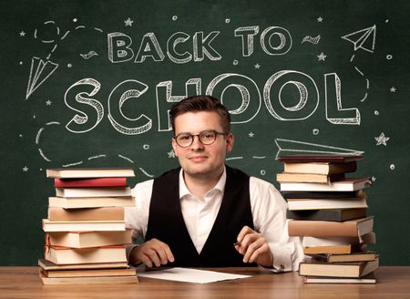 A young teacher in glasses sitting at classroom desk with pile of books in front of blackboard saying back to school drawing concept.