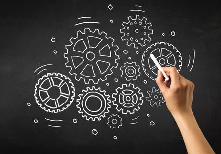 Female hand holding white chalk in front of a blackboard with gears drawn on it