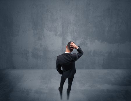 A young businessman with briefcase standing in blank empty space facing a grey urban wall, scratching his head concept.