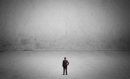 A tiny elegant businessman standing in large empty urban space with concrete walls and grey background concept