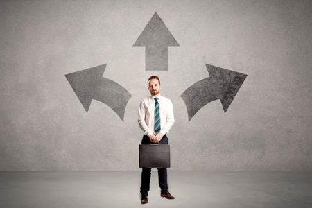 Charming businessman in doubt, choosing from three directions in front of a grey wall
