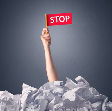 Female hand emerging from crumpled paper pile holding a red flag with stop written on it 