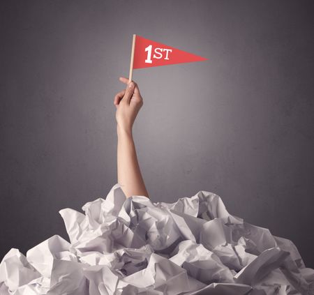 Female hand emerging from crumpled paper pile holding a red flag with first written on it