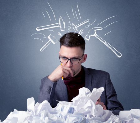 Young businessman sitting behind crumpled paper with drawn hammers hitting his head 