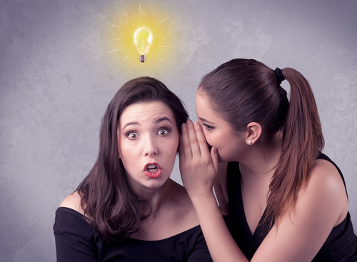 A young girl has an idea illustrated with a drawn glowing light bulb above the head, while a friend whispers a secret in her ear concept.