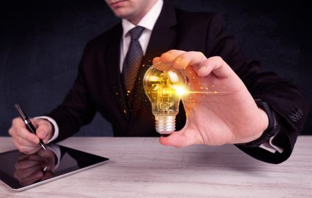 An elegant office worker holding a yellow sparkling light bulb in his hand while working in front of dark blue background concept.