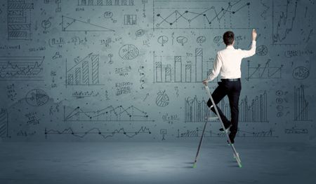A businessman in modern stylish elegant suit standing on a small ladder and drawing pie and block charts on grey wall background with exponential progressing  lines, circles, angles, blocks, numbers