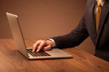 An office worker in elegant suit sitting at desk, typing on portable laptop with empty brown wall background