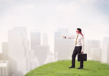 Young blindfolded businessman steps on a a patch of grass with a city in the background