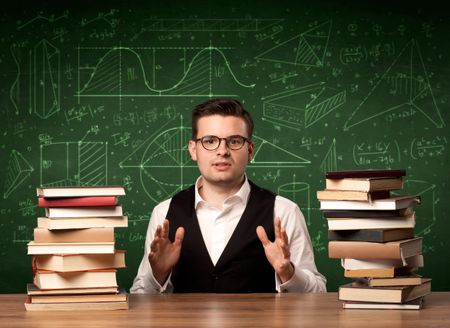 A young passionate male teacher sitting at school desk, reading a book, with area algorythm calculations and numbers on the blackboard concept.