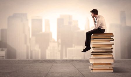 A serious student in elegant suit sitting on a pile of books looking over a brown sepia city landscape