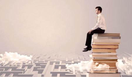 A serious businessman in suit sitting on a pile of giant books in front of a grey wall with clouds, labirynth