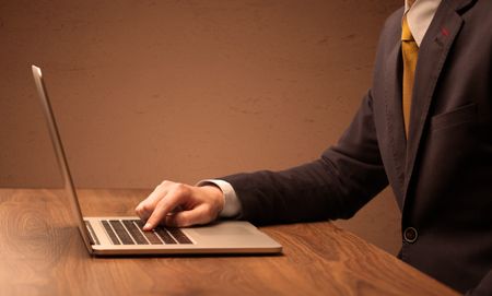 An office worker in elegant suit sitting at desk, typing on portable laptop with empty brown wall background