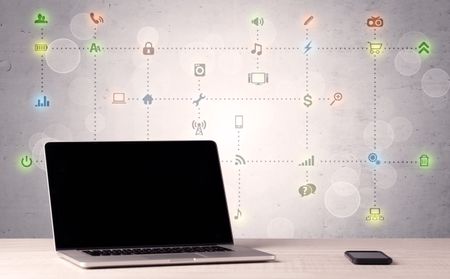 Notebook with blank screen sitting on office desk in front of background wall full of communication icons and illustrated connections concept