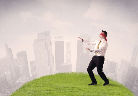 Young blindfolded businessman steps on a a patch of grass with a cloudy city in the background