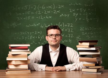 A young ambitious teacher in glasses sitting at classroom desk with pile of books in front of blackboard full of math calculations, numbers, back to school concept.