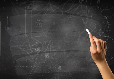 Female hand holding white chalk in front of a blackboard with scribbles and plans drawn on it