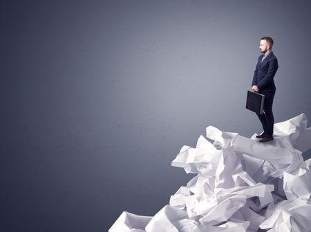 Thoughtful young businessman standing on a pile of crumpled paper with a dark grey background