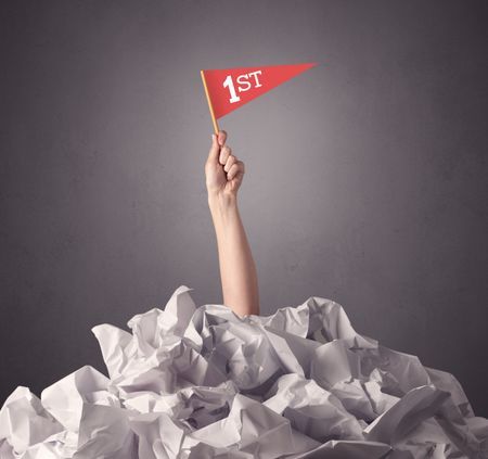 Female hand emerging from crumpled paper pile holding a red flag with first written on it