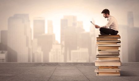 A serious businessman with laptop tablet in elegant suit sitting on a stack of books in front of cityscape