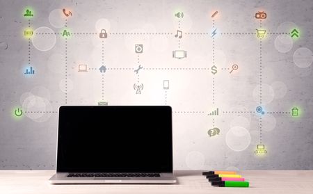 Notebook with blank screen sitting on office desk in front of background wall full of communication icons and illustrated connections concept