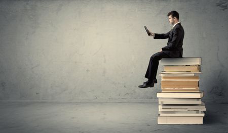 A serious businessman with tablet in hand in suit sitting on a pile of giant books in front of a textured grey wall.