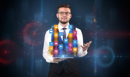 Elegant businessman holding tablet with hologram application icons and symbols above
