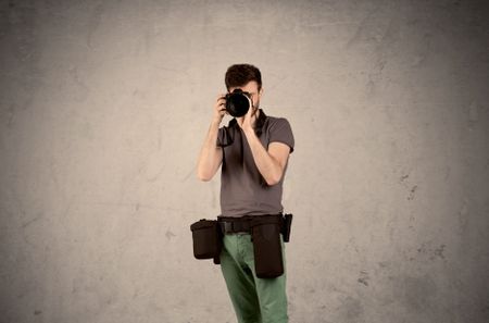 A professional male photographer with belt holding a camera and taking photos in front of clear grey urban wall background concept