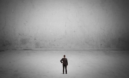 A tiny elegant businessman standing in large empty urban space with concrete walls and grey background concept