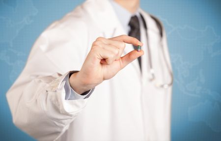 A male doctor in white coat in front of a blue wall, with a stethoscope on one shoulder holding a pill between his fingers