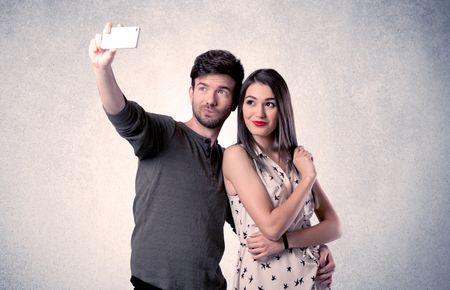 A young couple in love taking selfie with a mobile phone in the handsome guy's hand in front of an empty clear grey wall background concept