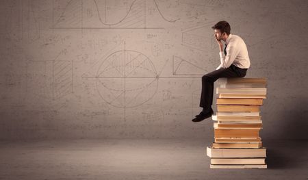 A serious businessman in suit sitting on a pile of giant books in front of a greyish brown wall with drawn lines, angles, numbers, circles and curves.