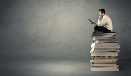 A serious businessman with laptop tablet in elegant suit sitting on a stack of books in front of dark grey background