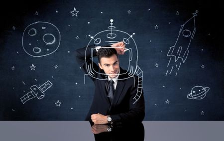 A young happy businessman in elegant suit drawing helmet, satellite and rocket in empty space with a chalk illustration concept