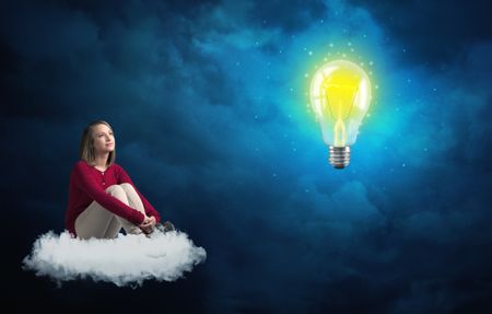 Caucasian woman sitting on a white fluffy cloud looking and wondering at a big, bright, shiny, glowing yellow lightbulb