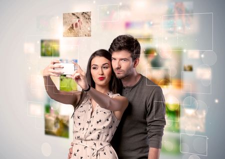 Collecting memories concept with photos in backbround and a happy young couple in love taking selfie with a mobile phone