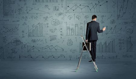 A businessman in modern stylish elegant suit standing on a small ladder and drawing pie and block charts on grey wall background with exponential progressing curves, lines, circles,  blocks, numbers