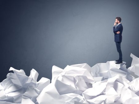 Thoughtful young businessman standing on a pile of crumpled paper with a blueish grey background