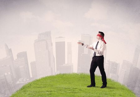 Young blindfolded businessman steps on a a patch of grass with a cloudy city in the background