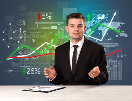 Young handsome businessman sitting at a desk with stocks and progress charts behind him