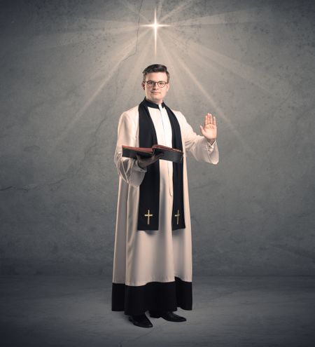 A young male priest in black and white giving his blessing in front of grey wall with glowing cross concept.