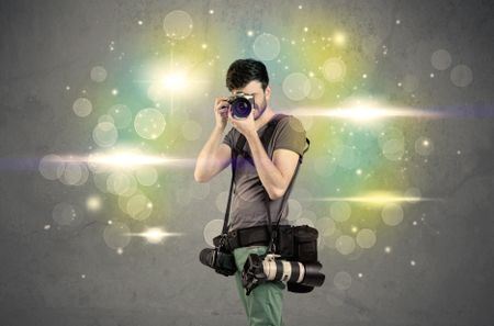 A young amateur photographer with professional camera equipment taking picture in front of grey wall full of colorful bokeh and glowing lights concept