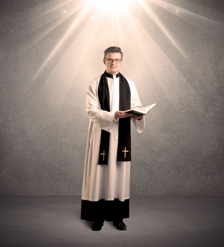 A male religious young priest in black and white dress giving his blessing, holding the holy bible while being illuminated from strong light beams coming from above concept