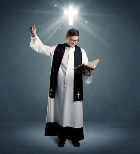 A young caucasian priest with deep faith blessing with the holy bible in his hand in front of blue wall background with illustrated glowing cross concept.