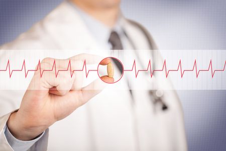 A male doctor in white coat with a stethoscope on one shoulder holding a pill between his fingers focused on a heartbeat graph