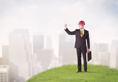 A cheerful caucasian elegant construction manager standing in small green grass and drawing with a pen in front of faded city landscape, tall buildings concept.