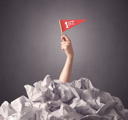 Female hand emerging from crumpled paper pile holding a red flag with first written on it