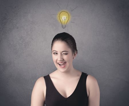 A teen student girl with funny facial expression has a good idea illustrated by a drawn light bulb lighting up above the head on the grey wall background concept.