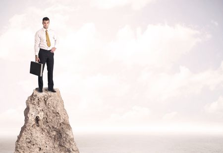 Successful sales person with brief case standing on top of a mountain cliff edge looking above the landscape between the clouds